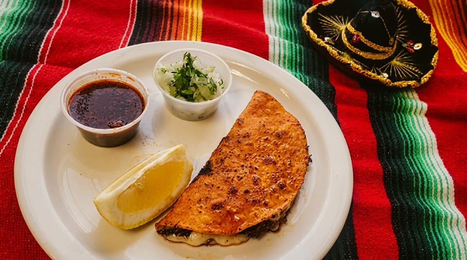 A birria quesadilla with consome, onions, cilantro, and a lemon wedge on a colorful striped blanket.