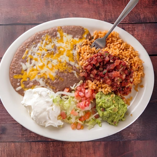 A white oval plate with refried beans, rice, pico de gallo, sour cream, lettuce, tomatoes, and guacamole.