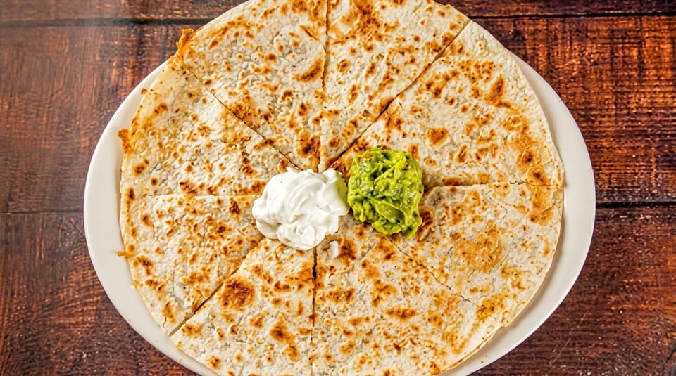 A round quesadilla, cut into eight slices, with dollops of sour cream and guacamole on a white plate.