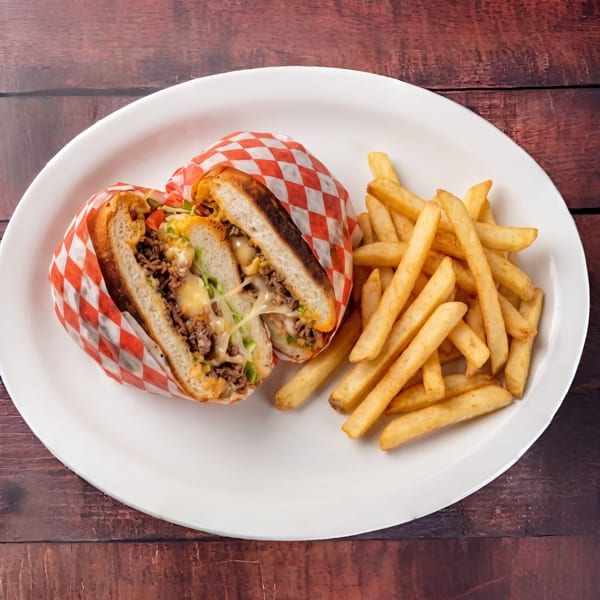 A sliced cheesesteak sandwich wrapped in checkered paper, served with golden french fries on a white plate.